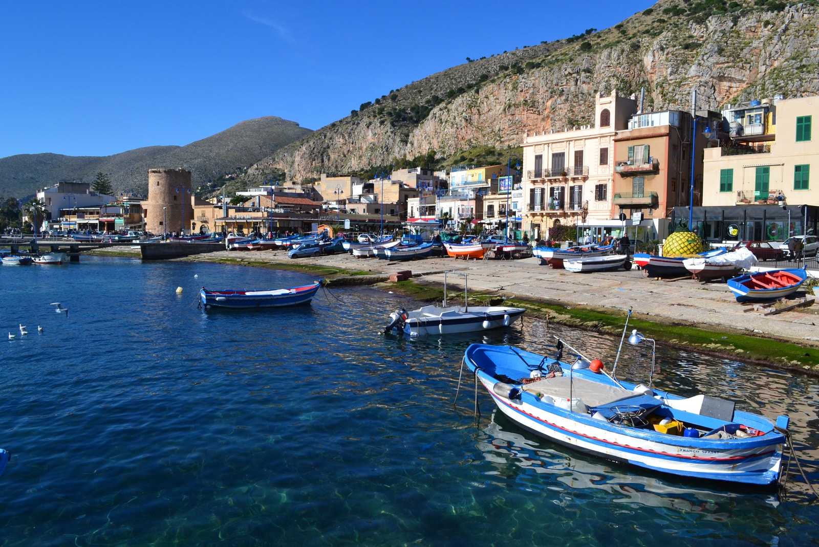 The historic Tonnara watchtower in Mondello harbor, with the limestone cliffs of Monte Gallo rising dramatically in the background.