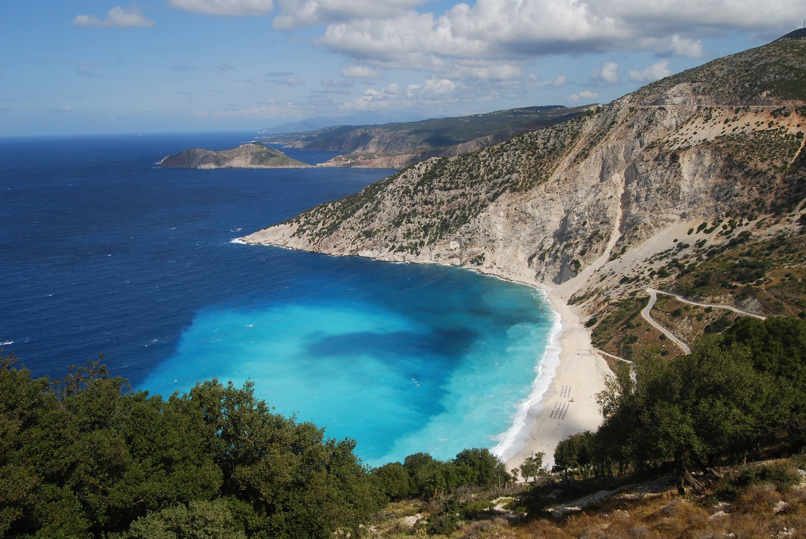 An elevated view of the curved Myrtos Beach coastline, showing the contrast between the white pebbles, electric blue water, and the winding road carved into the mountainside.