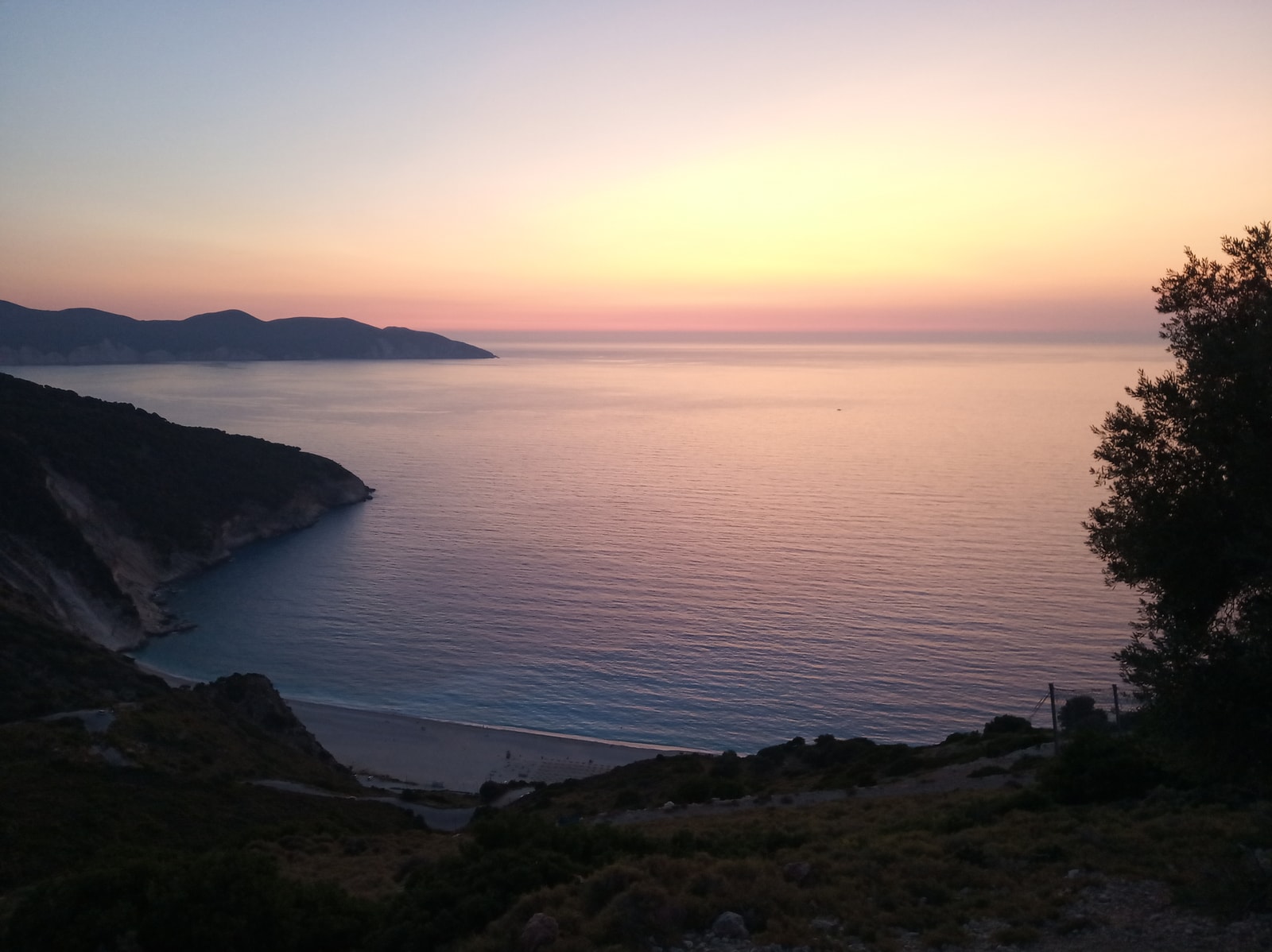 A view of Myrtos beach at sunset with soft pink and lavender colors in the sky, seen from the hills leading up to Divarata.