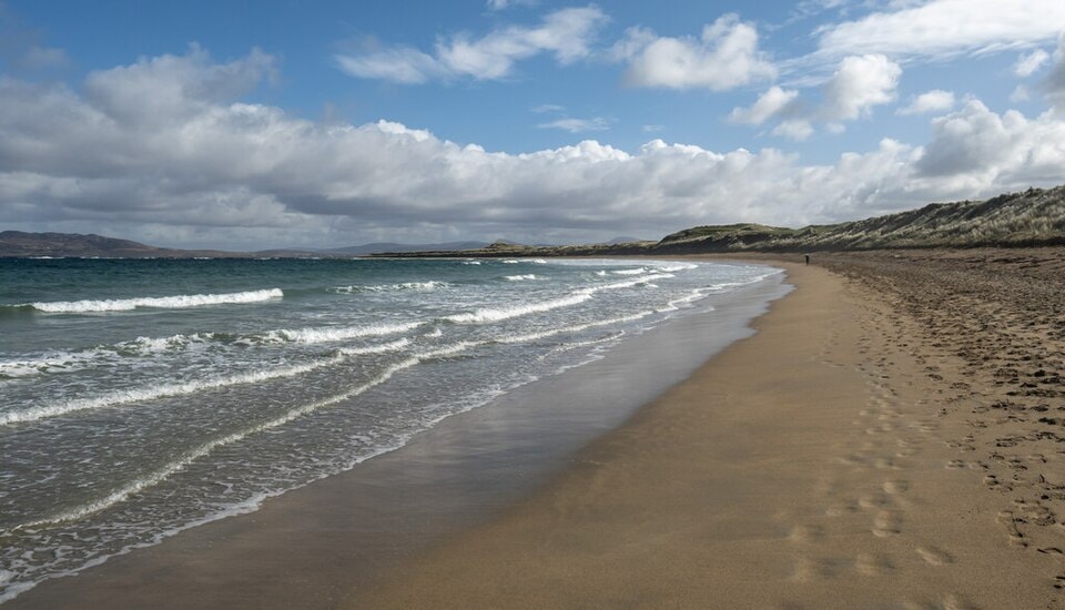 A dramatic beach scene with wet, reflective sand in the foreground, gentle waves breaking, and a sky filled with dynamic, moody clouds.