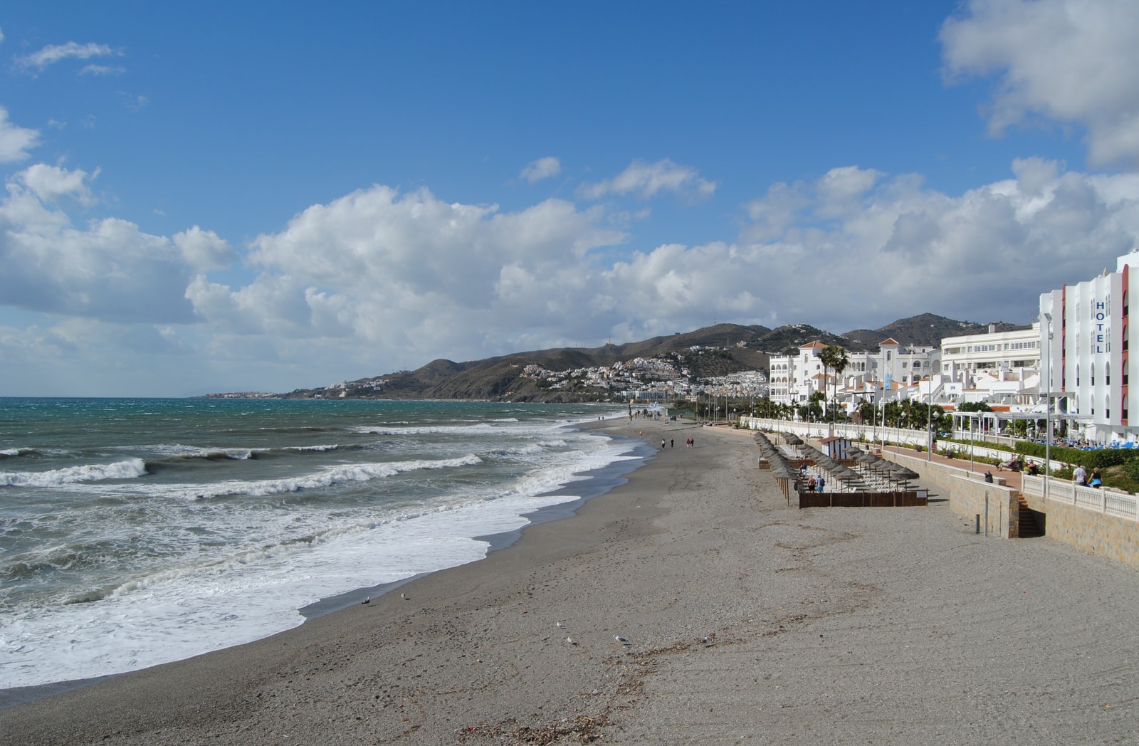 A long, pebbly shoreline curves along the coast, with a promenade, buildings, and dramatic clouds in the sky.