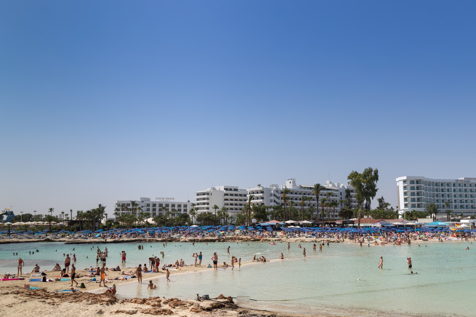 A wide view of Nissi Beach shows families relaxing on the sand and swimming, with large resort hotels in the background.