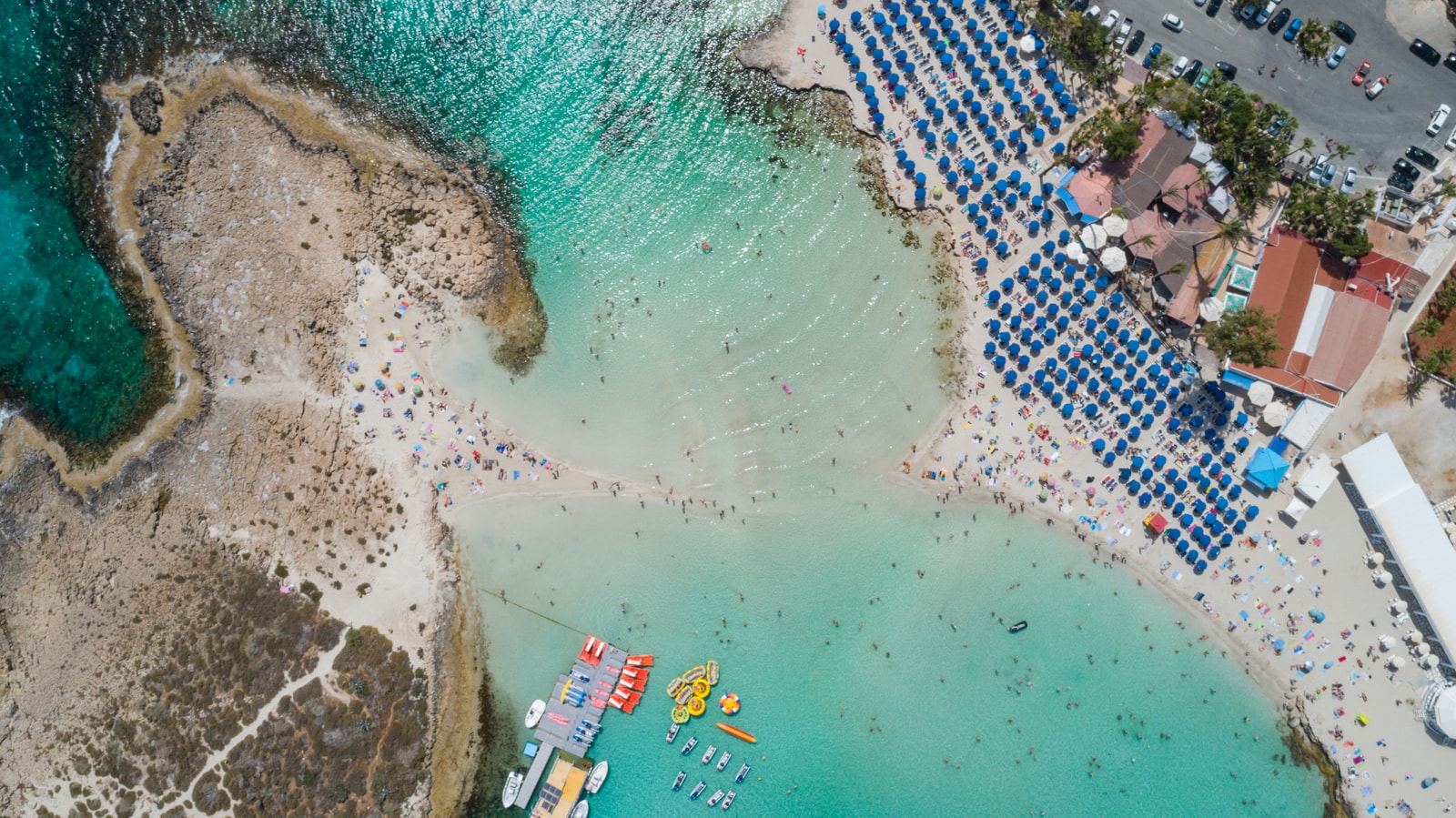 An aerial view from directly above Nissi Beach shows the thin sandbar connecting the mainland to the islet, with people wading across.