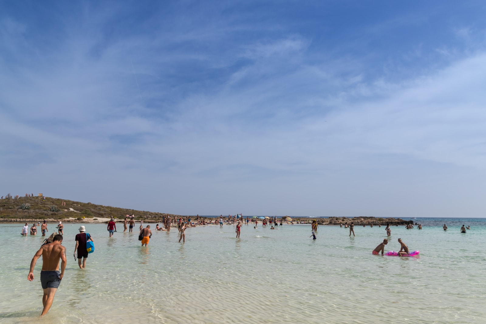 A ground-level view of tourists wading through the knee-deep, bright turquoise water over the sandspit toward the islet.