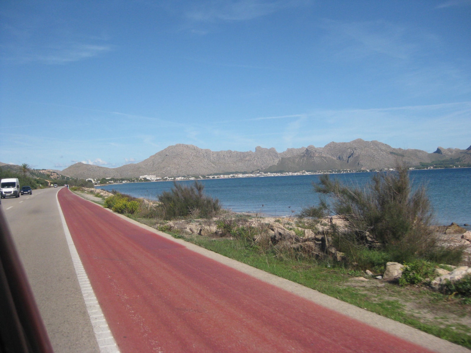 A paved cycle path runs alongside a bay with calm blue water, with a mountainous peninsula visible in the distance.