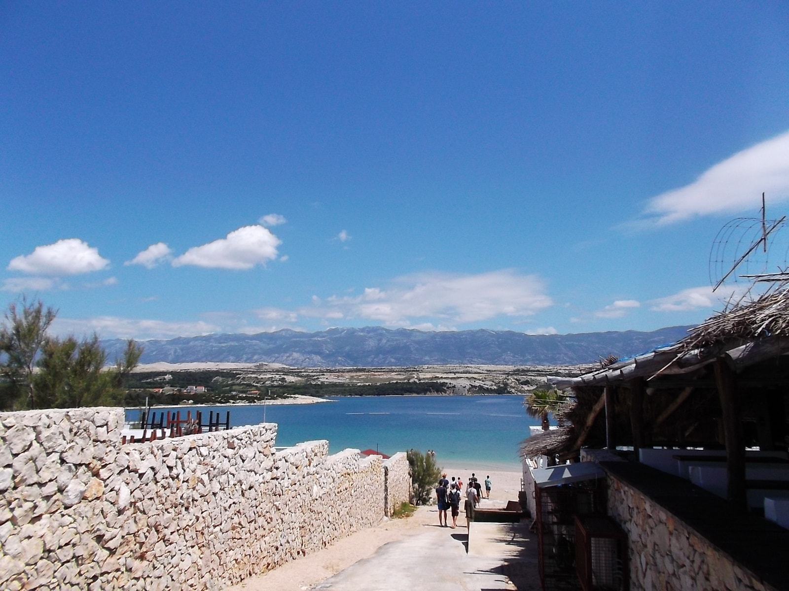 A stone pathway framed by a traditional dry-stone wall leads down to a sparkling turquoise bay in Novalja.