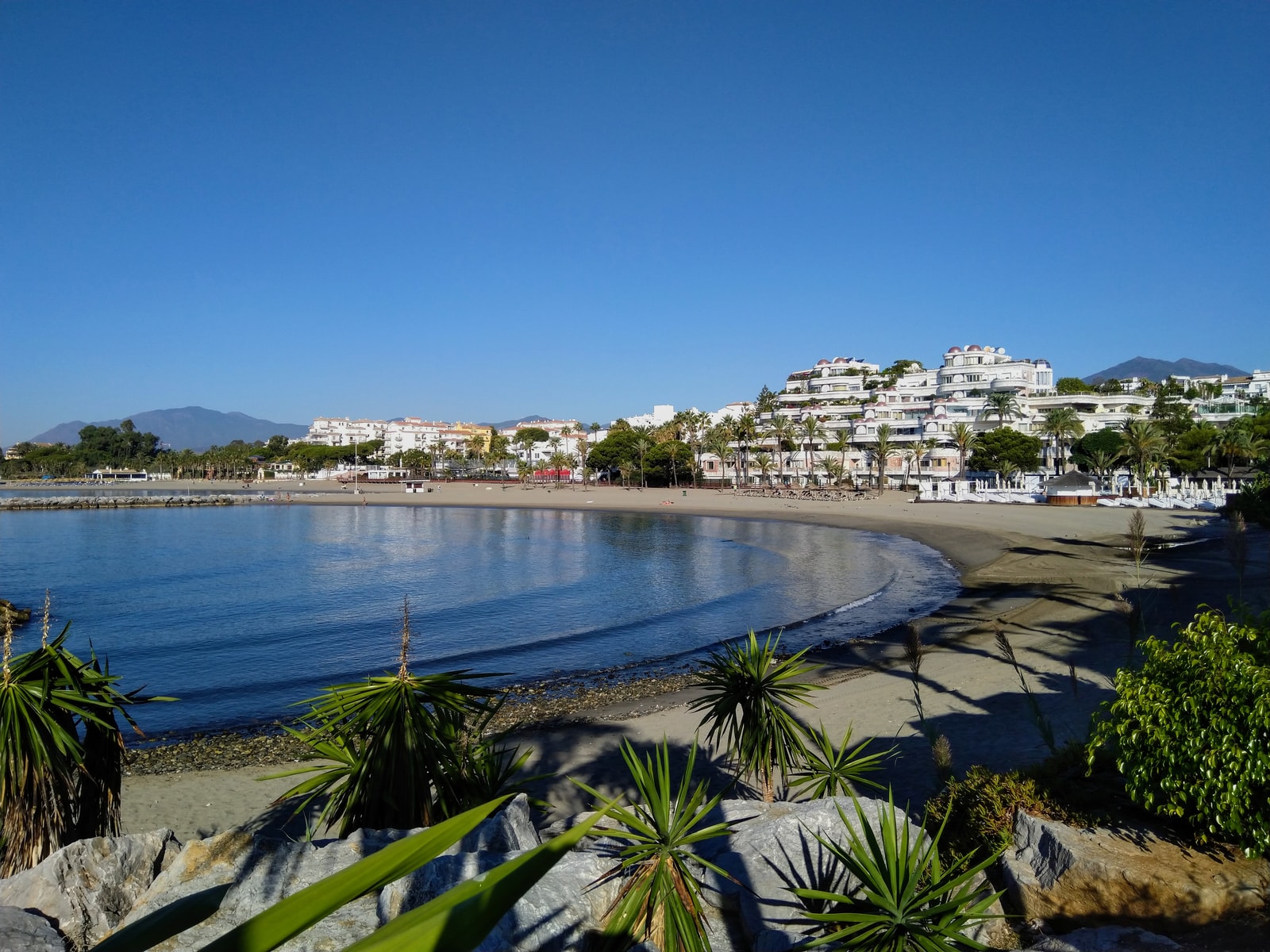 A crescent-shaped cove beach in Nueva Andalucía, created by a stone breakwater, with luxury hillside apartments overlooking the sea.