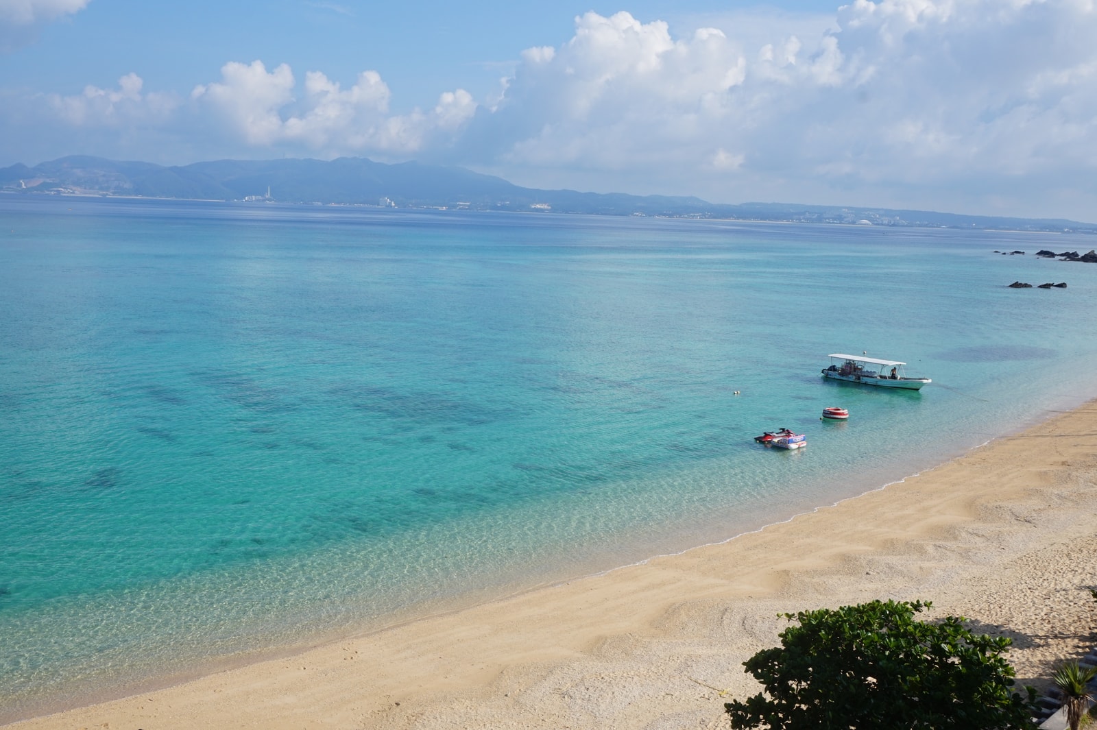 A wide shot of a calm turquoise sea with small boats and mountains in the distance.