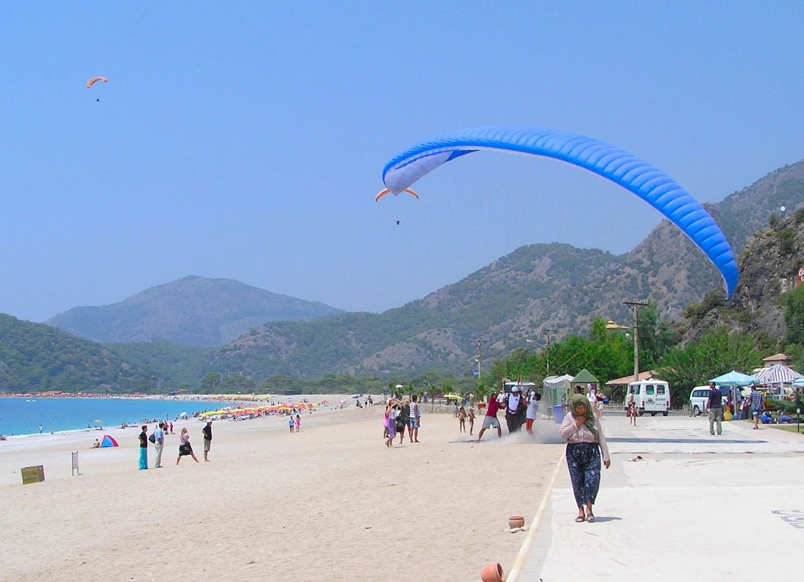 A paraglider with a red and white canopy floats above a bustling beach promenade with sun umbrellas and loungers.