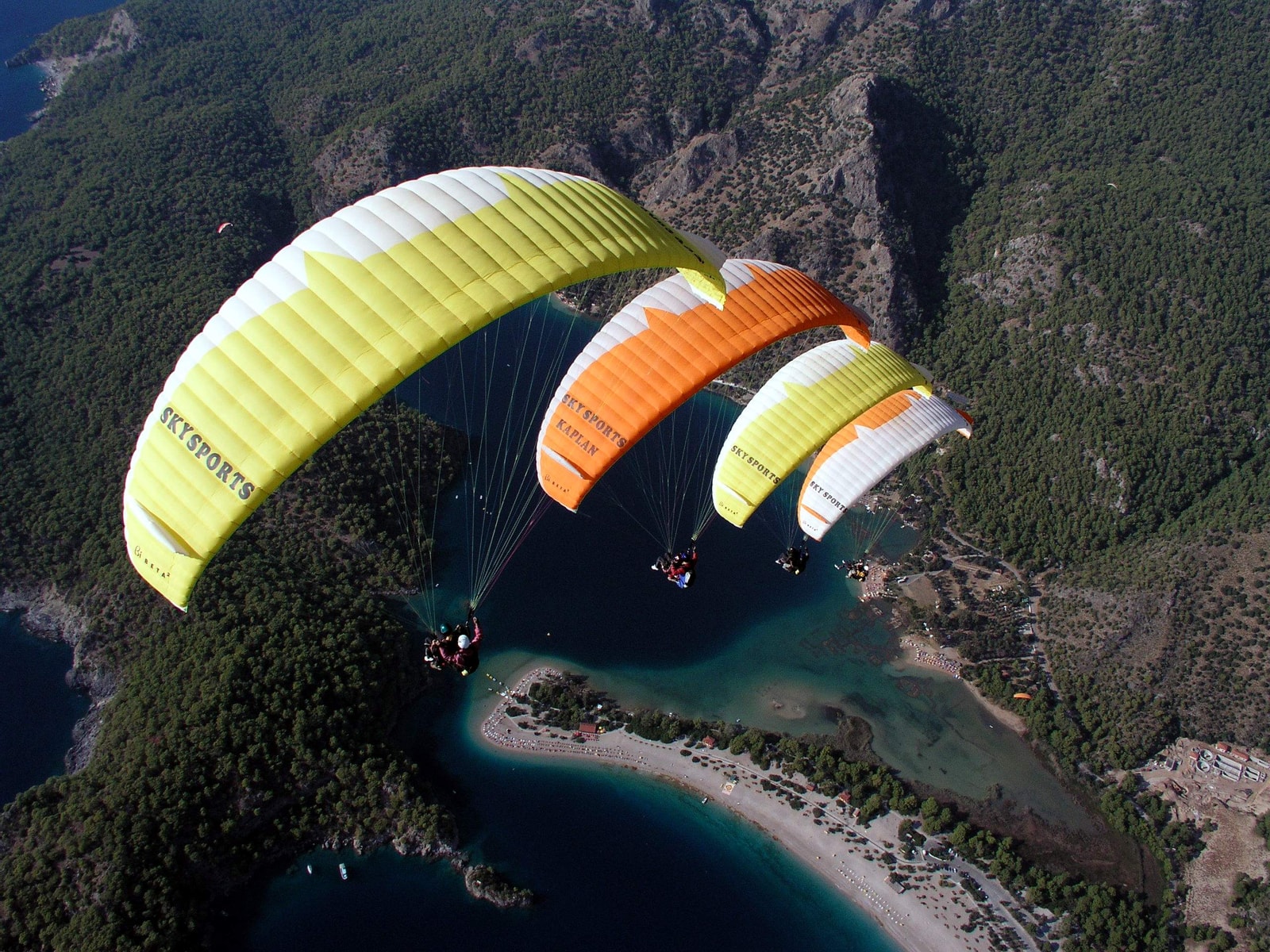 Three colorful paragliders soar high above the turquoise Blue Lagoon and the green, mountainous coastline.