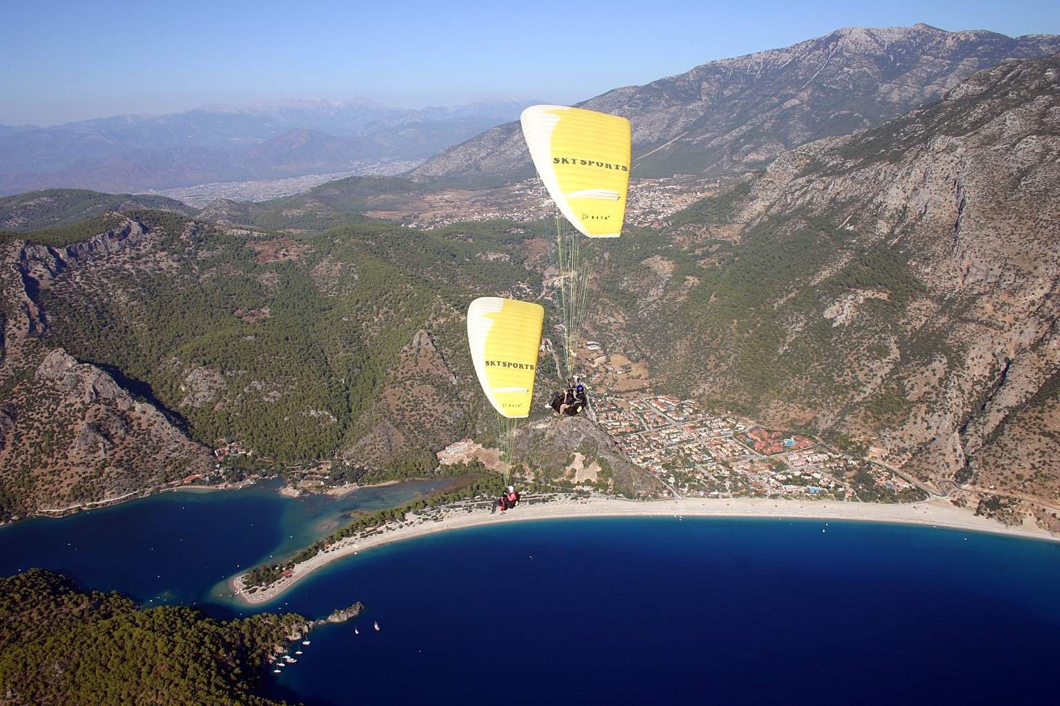 Two paragliders with yellow canopies fly over the bay, with the resort town and beach visible below.