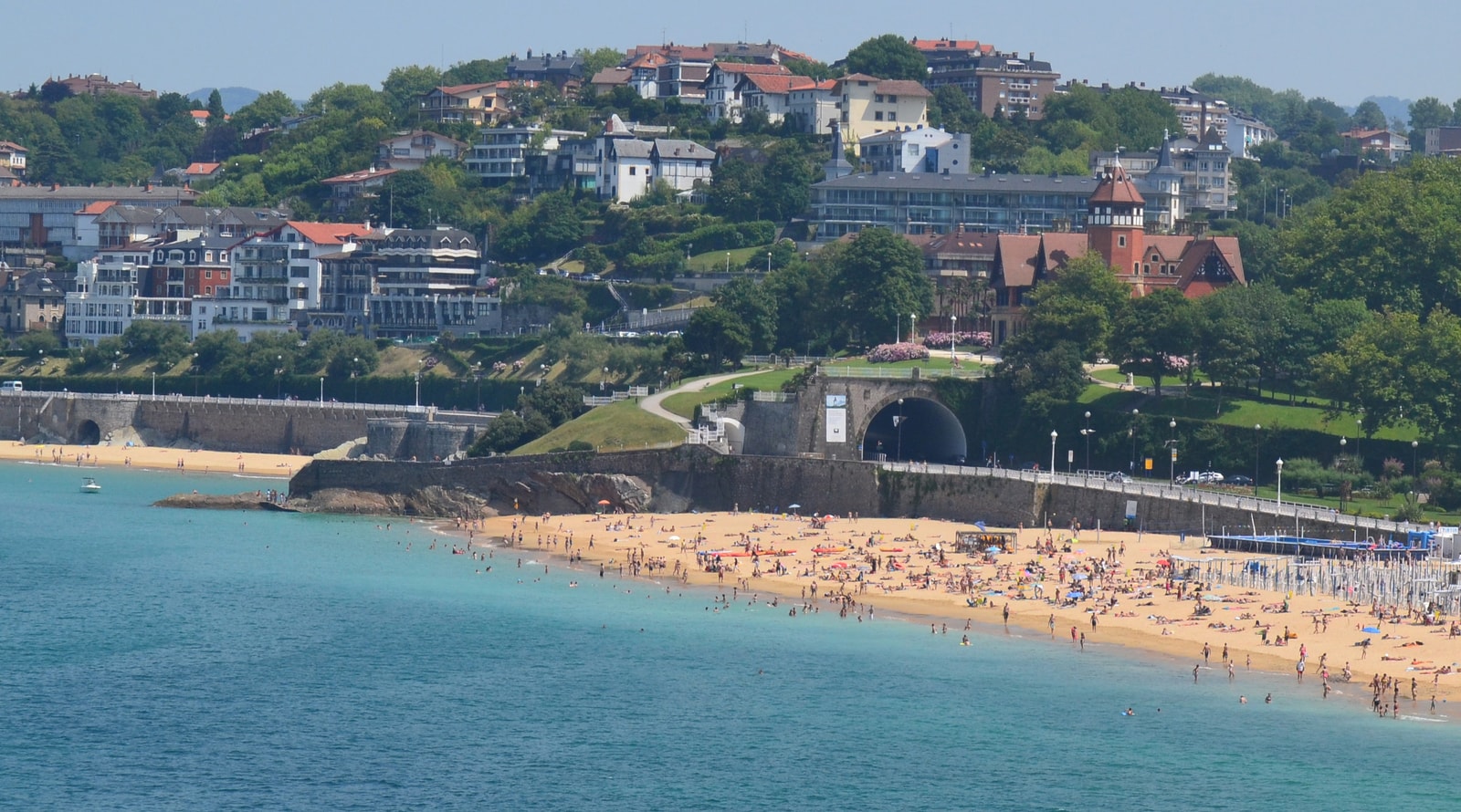 A view of Ondarreta Beach filled with colorful umbrellas, with the Loretopea coastal tunnel to the left and Miramar Palace on the hill above.