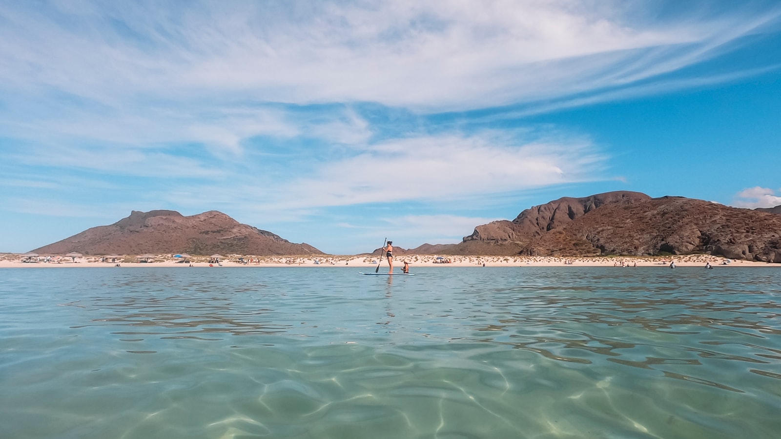 A person on a paddleboard glides across calm, transparent turquoise water near a desert coastline.
