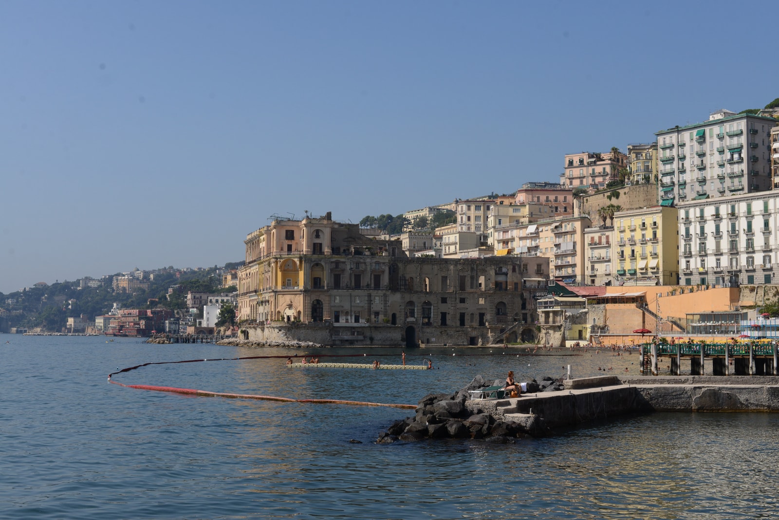 A view of the historic Bagno Elena beach club, with swimmers on the sandy shore and the massive, unfinished Baroque Palazzo Donn'Anna rising out of the sea beside it.