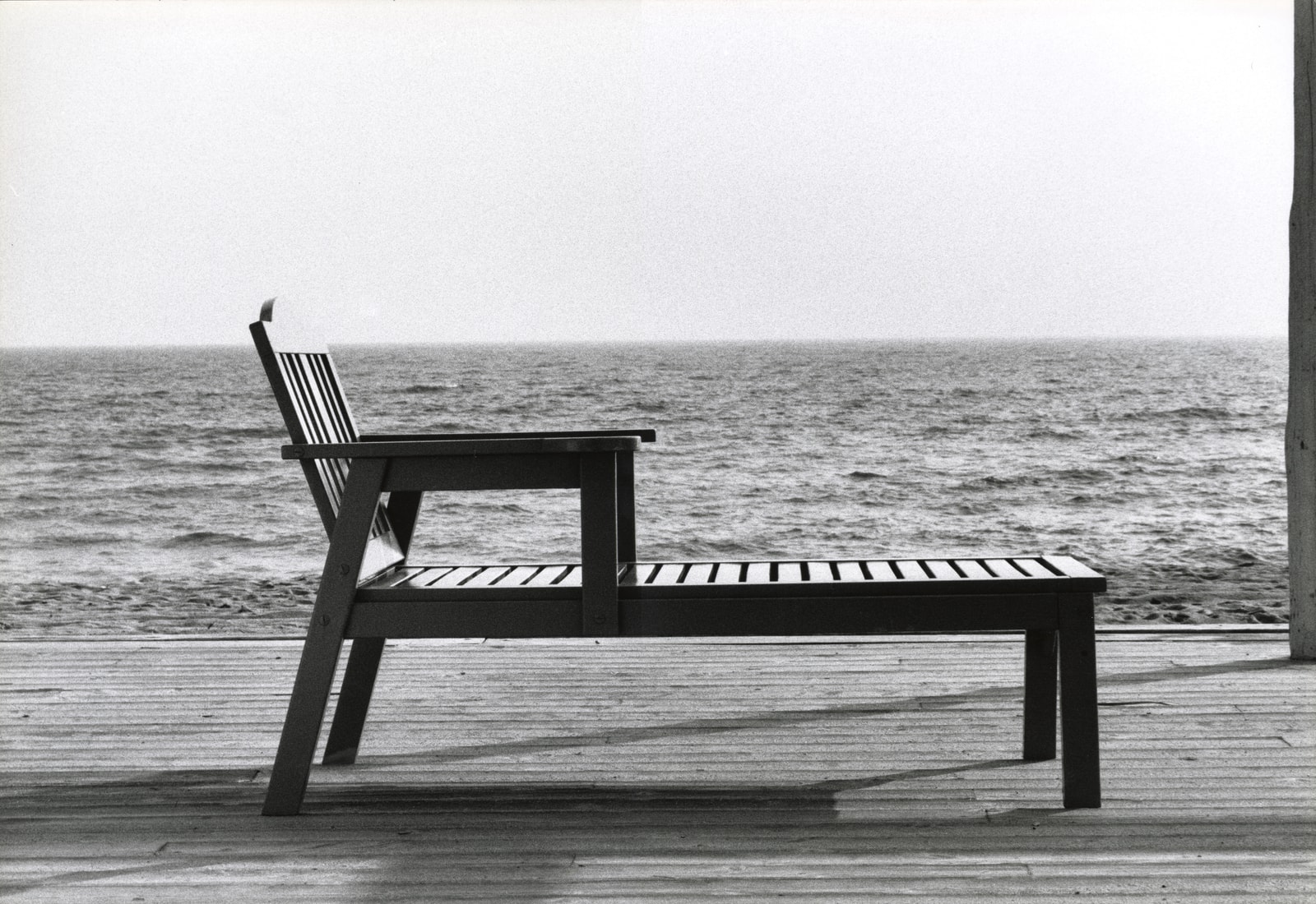 A minimalist black-and-white photo of a modern wooden chaise longue on the deck of a beach club.