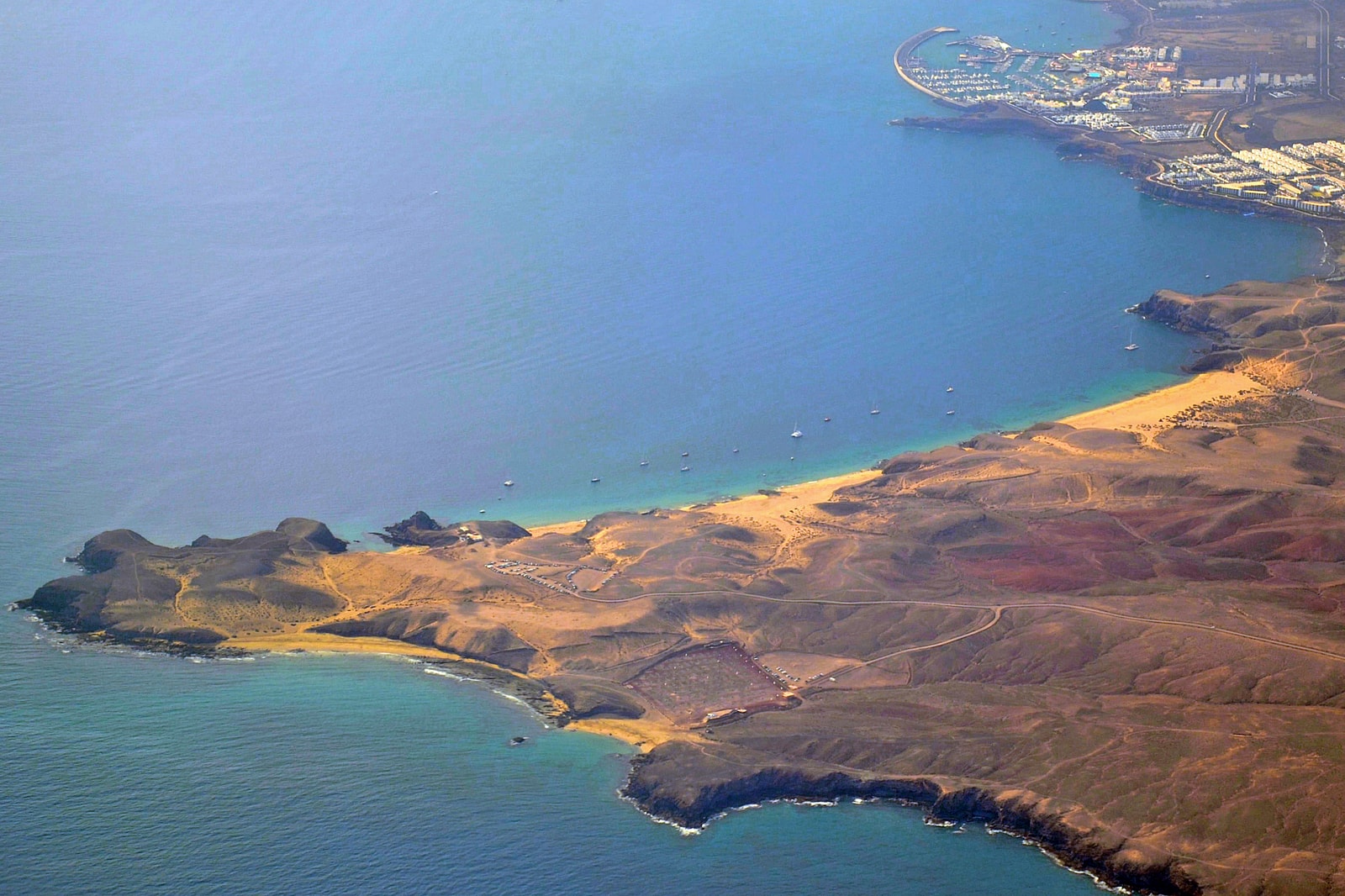 An aerial view shows a series of golden sand coves separated by dark rocky outcrops, with clear blue water.