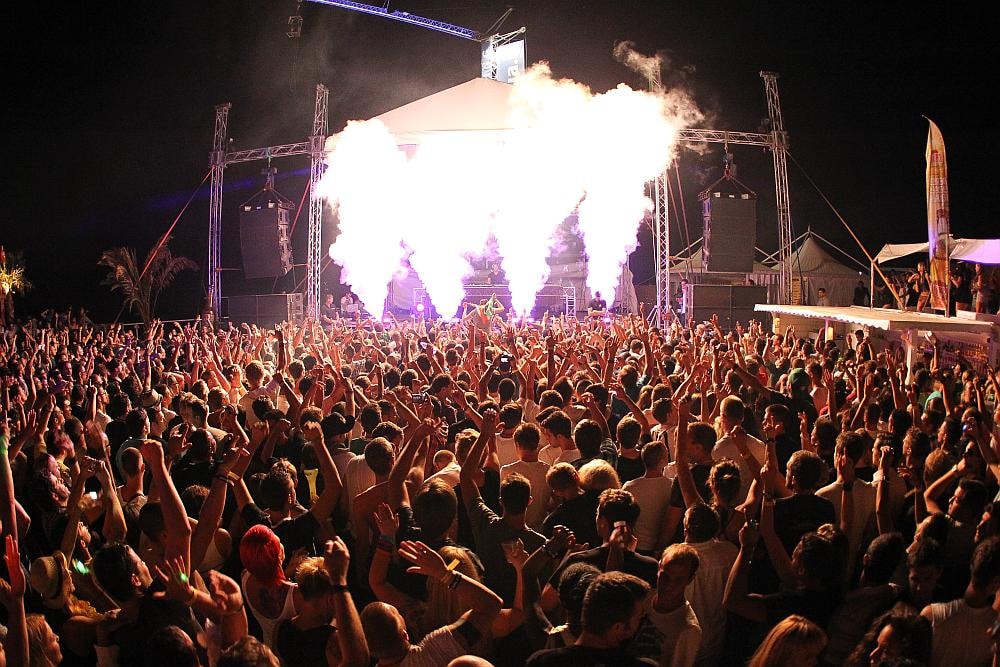 A large, energetic crowd dances at Papaya Club under vibrant stage lights and pyrotechnic smoke.