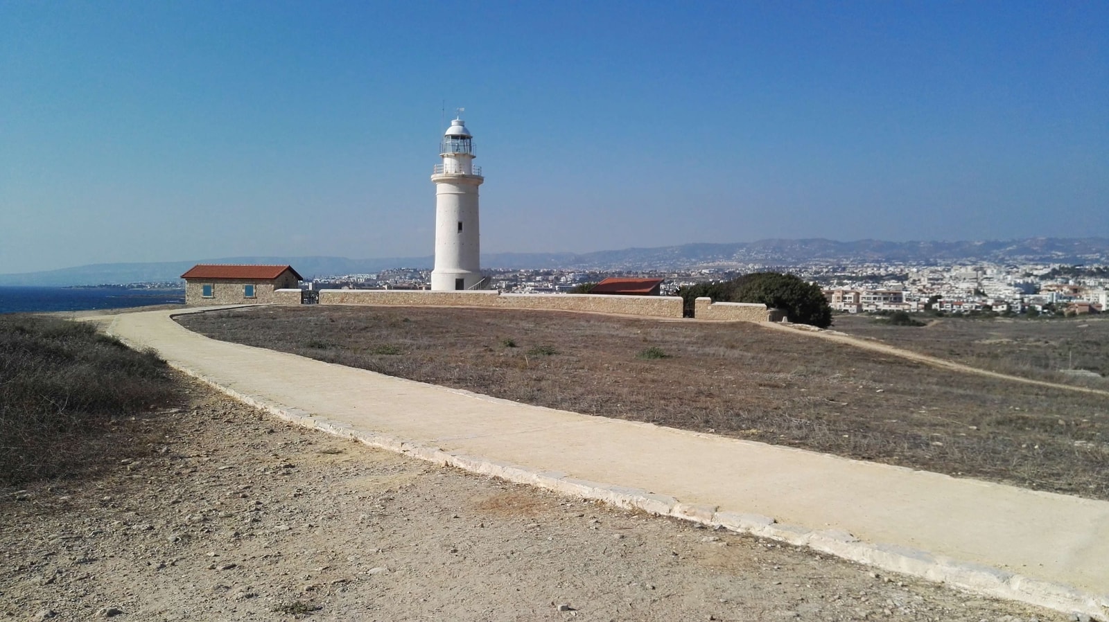 A view of the Paphos lighthouse, a coastal path, and the sea from within the archaeological park.