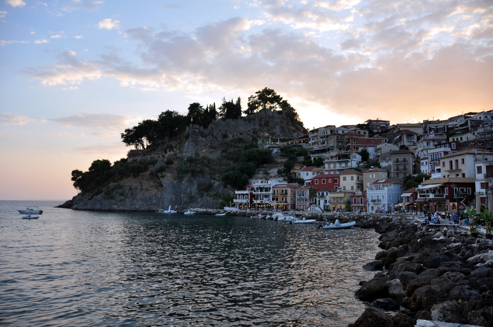 A scenic view of Parga's harbor at twilight, with the castle on the hill and lights starting to turn on in the town.