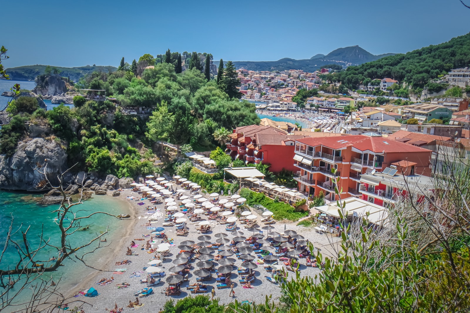 Rows of colorful beach umbrellas on Lichnos Beach, with resort buildings and green hills in the background.