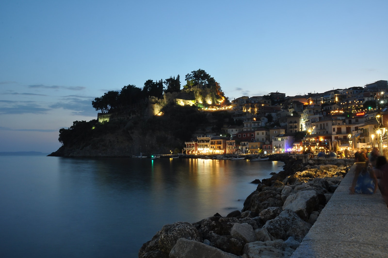 The waterfront of Parga illuminated at night, with lights reflecting on the calm harbor water and the castle lit up on the hill.