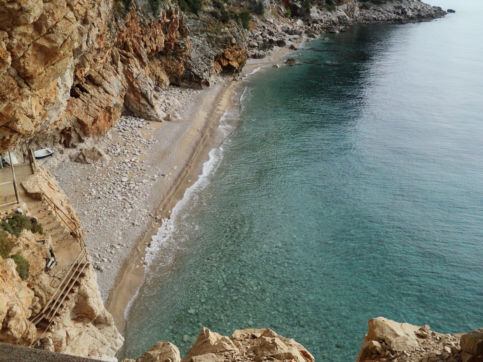 A view of Pasjača beach from the cliffside path, showing the stairs and railing leading down to the turquoise water.