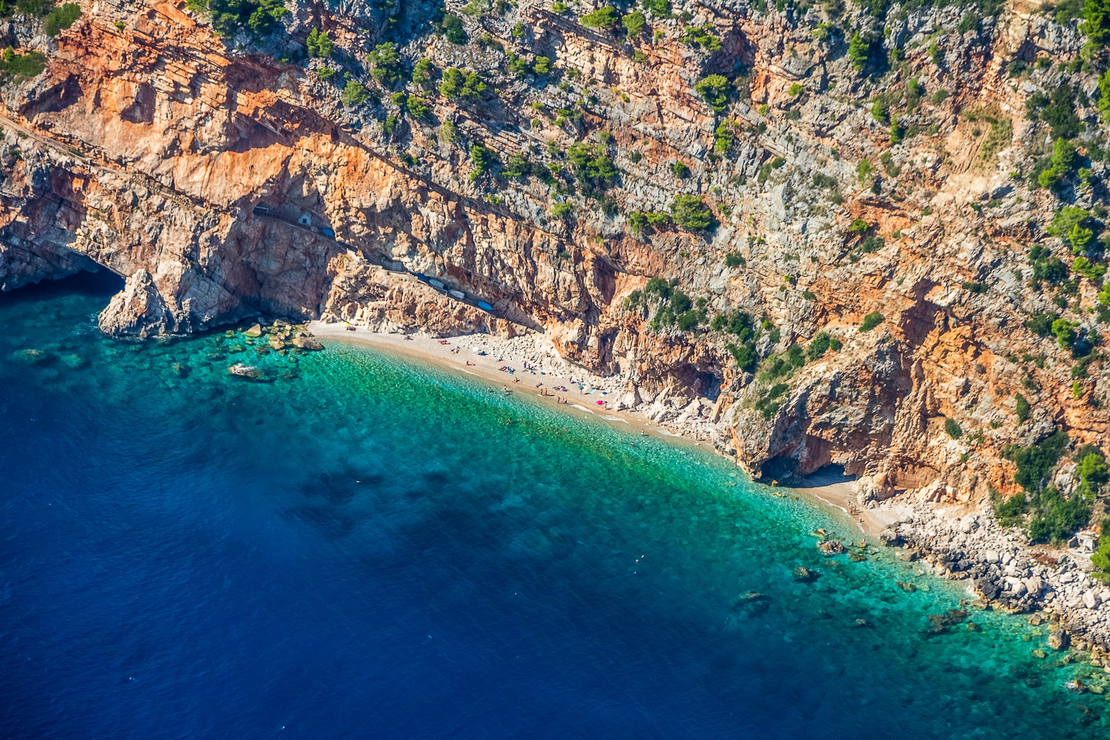 An aerial view of a narrow strip of beach at the bottom of immense, steep, rust-colored cliffs.