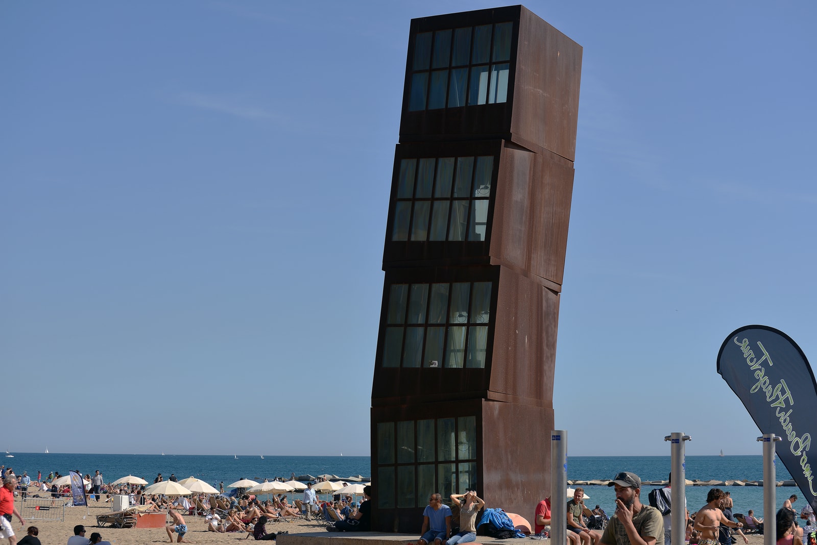 Pedestrians walk along the wide Passeig Marítim promenade, with palm trees on one side and the beach and a large sculpture on the other.