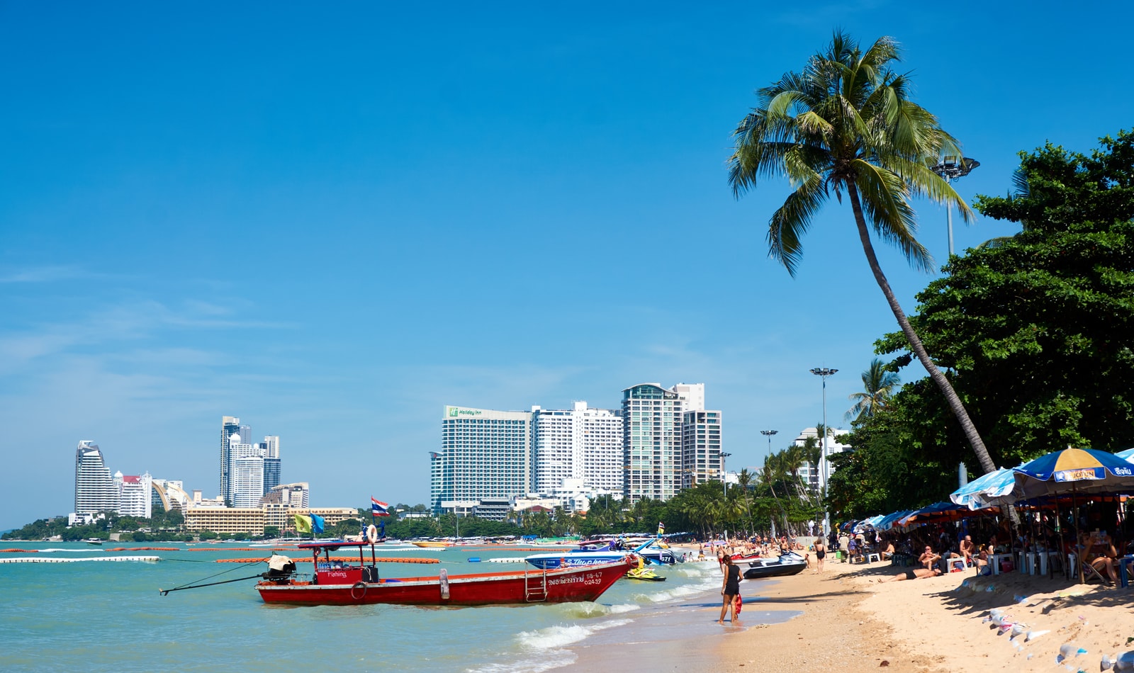 A vibrant beach scene at Pattaya with colorful umbrellas, palm trees, and tour boats moored in the shallow, turquoise water under a clear blue sky.