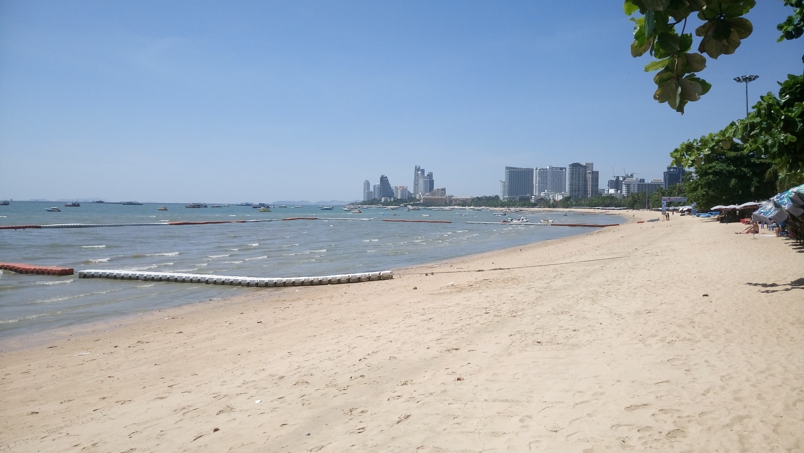 A sunny day at Pattaya beach, showing the wide sandy shore, the city skyline in the background, and buoys in the water.