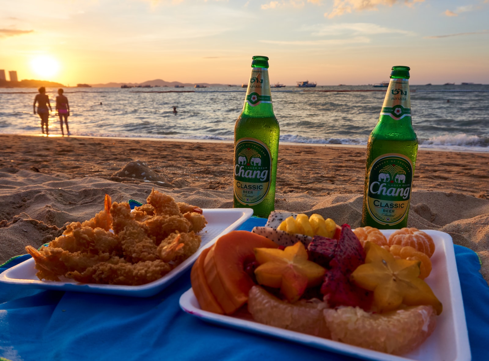 A casual beachside dining setup with food laid out on a mat, overlooking the sea as the sun sets in a warm, orange sky.