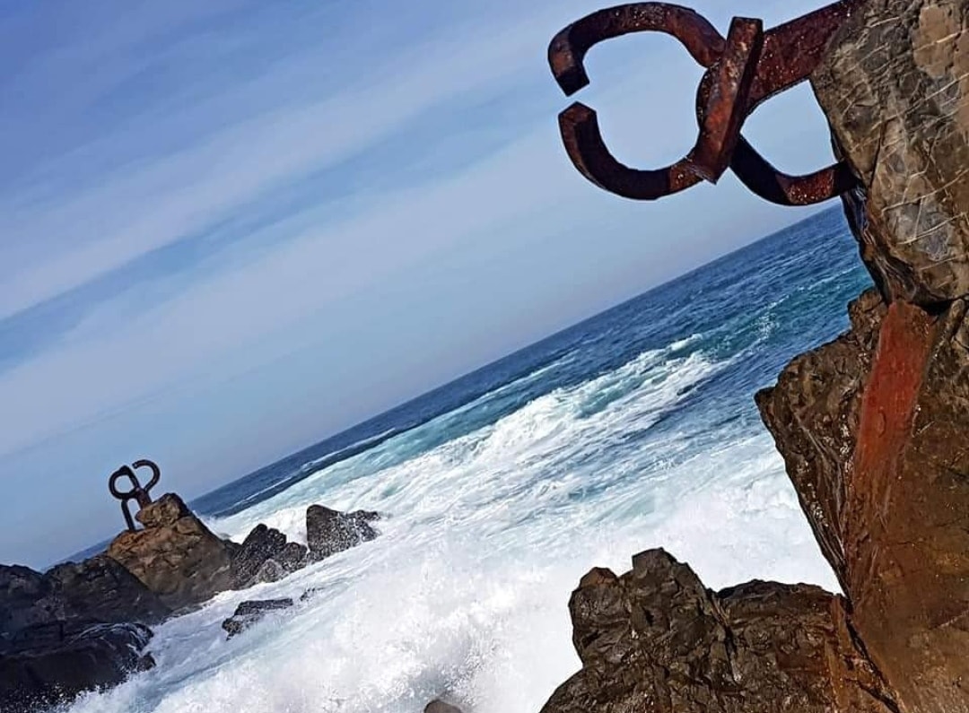 Three large, rust-colored steel sculptures resembling claws are anchored into rocks as waves crash around them.