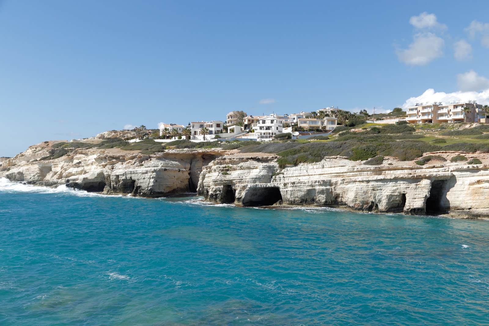 A panoramic view of pale limestone cliffs and sea cave openings meeting the bright blue sea under a sunny sky.