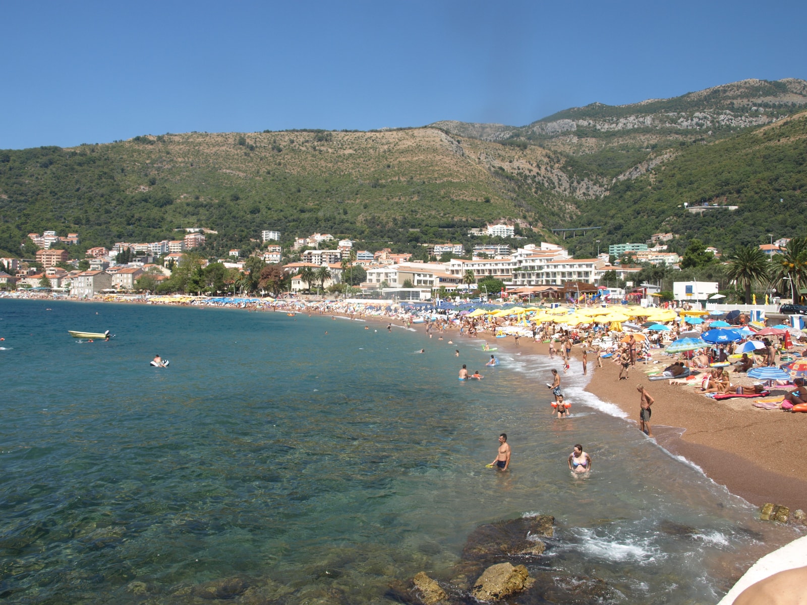 A view of a long pebble beach lined with umbrellas, with a town and green mountains in the background.