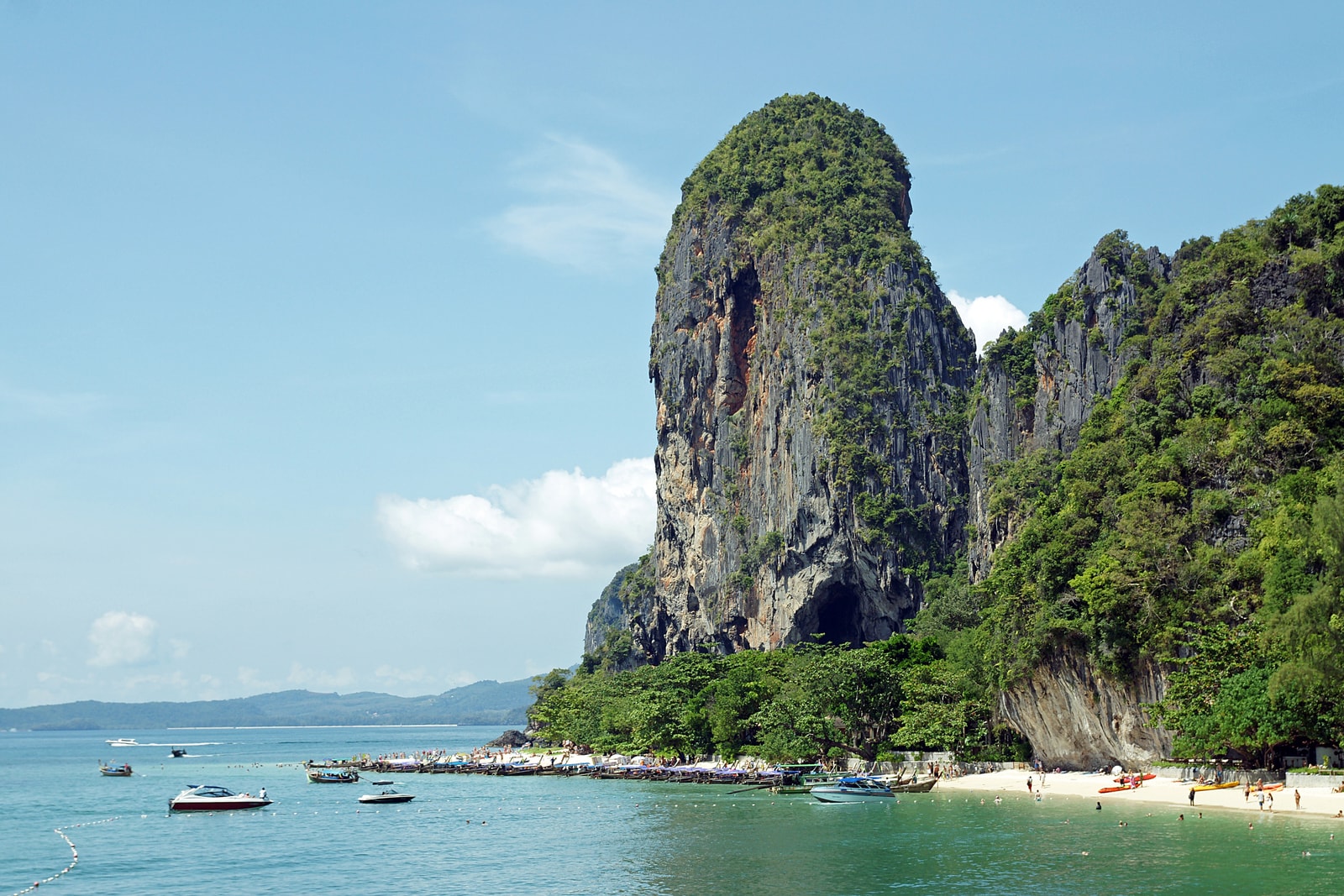 Longtail boats are moored along a sandy beach at the base of a towering, vegetation-covered limestone cliff.