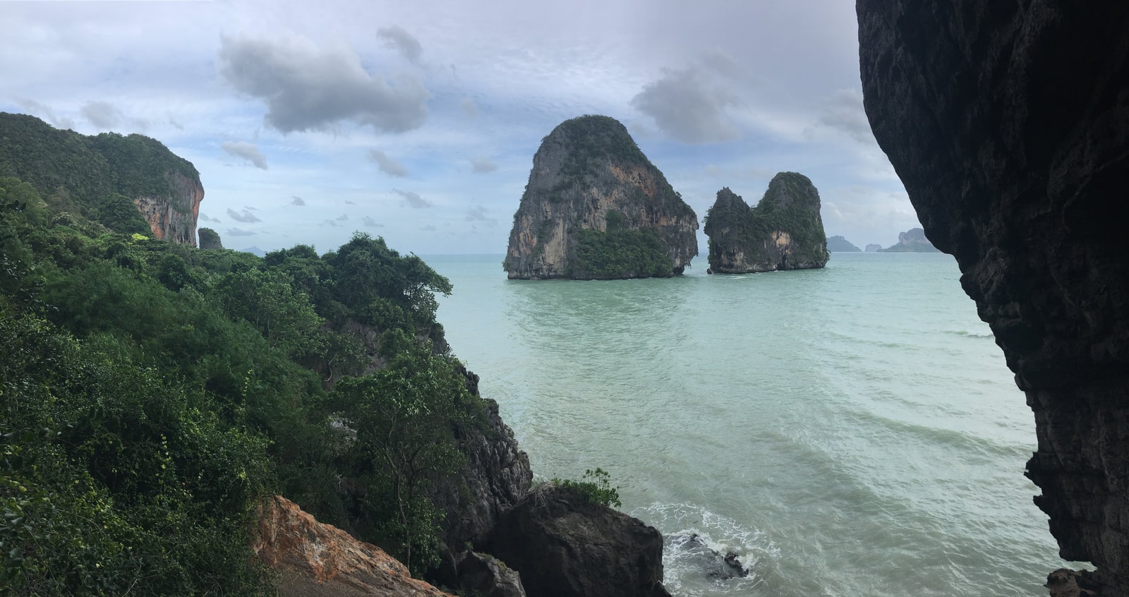 An elevated view from a cave looks out over Phra Nang Beach, showing the pale green water, tropical foliage, and distant limestone formations.
