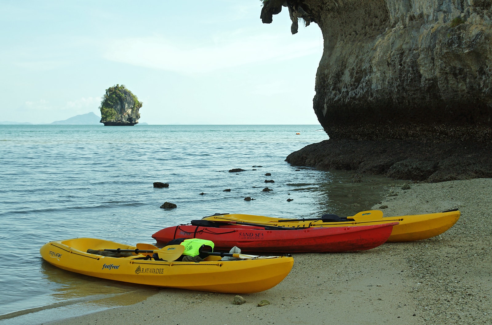 Colorful kayaks are lined up on the shore of Phra Nang Beach, with the massive limestone cliff overhang in the background.