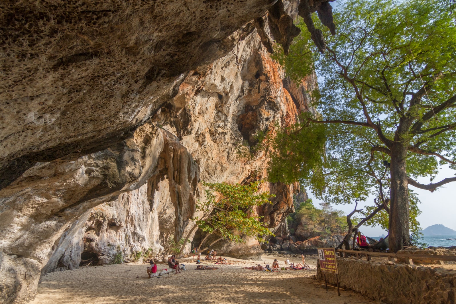 The entrance to the Princess Cave at Phra Nang Beach shows offerings within, while beachgoers relax on the sand under the massive rock overhang.