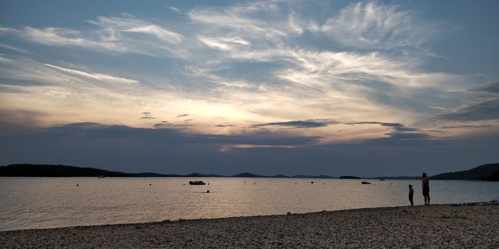 A wide-angle sunset over a quiet pebble beach in Pirovac, with silhouettes of people enjoying the view.