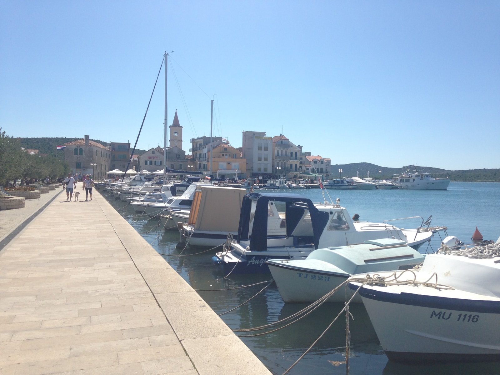 A scenic view of Pirovac's harbor, showing small boats docked along the stone promenade with the church tower in the background.