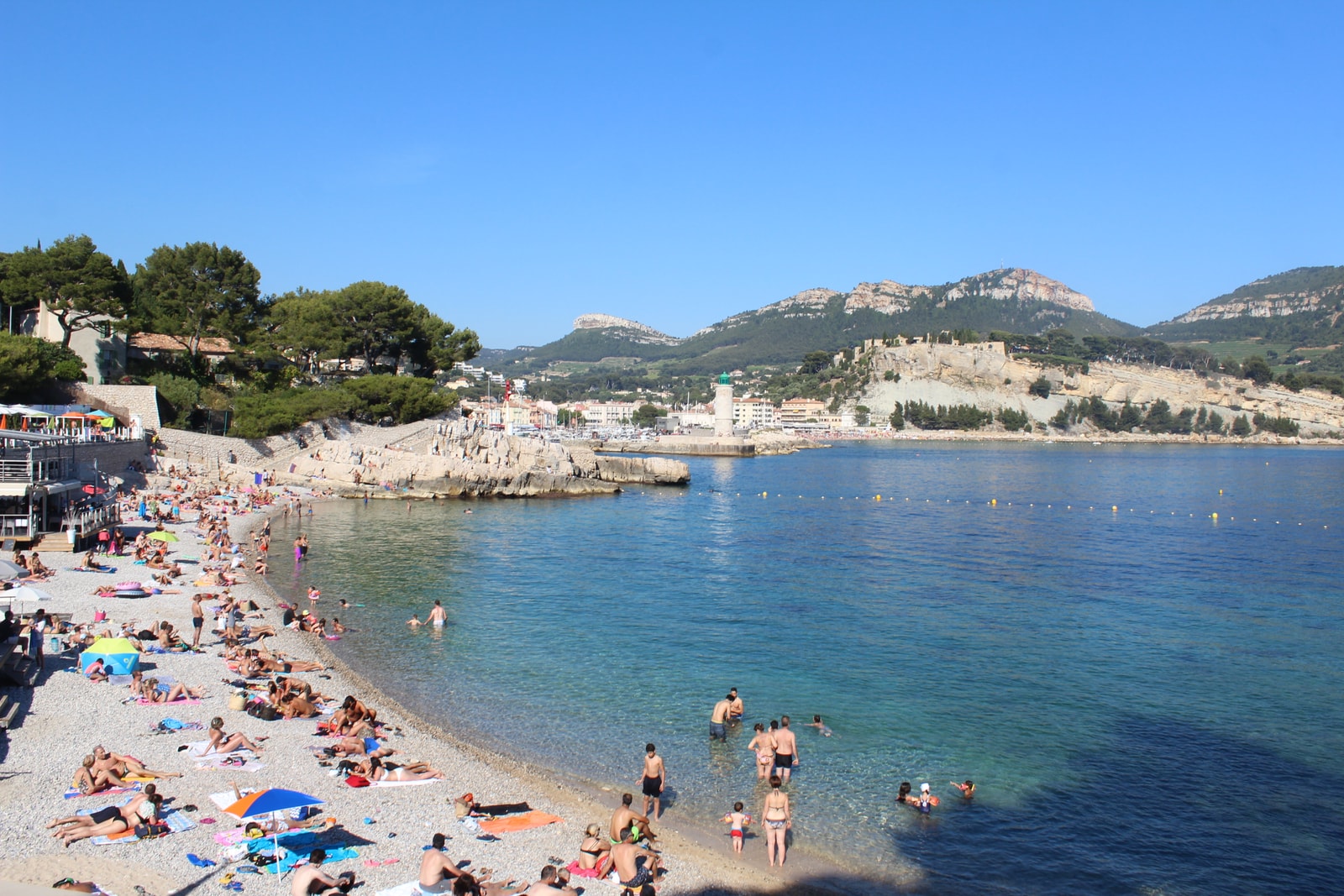 People relaxing and swimming at Plage de la Grande Mer in Cassis, with colorful umbrellas on the sand and the historic castle visible on a cliff in the background.