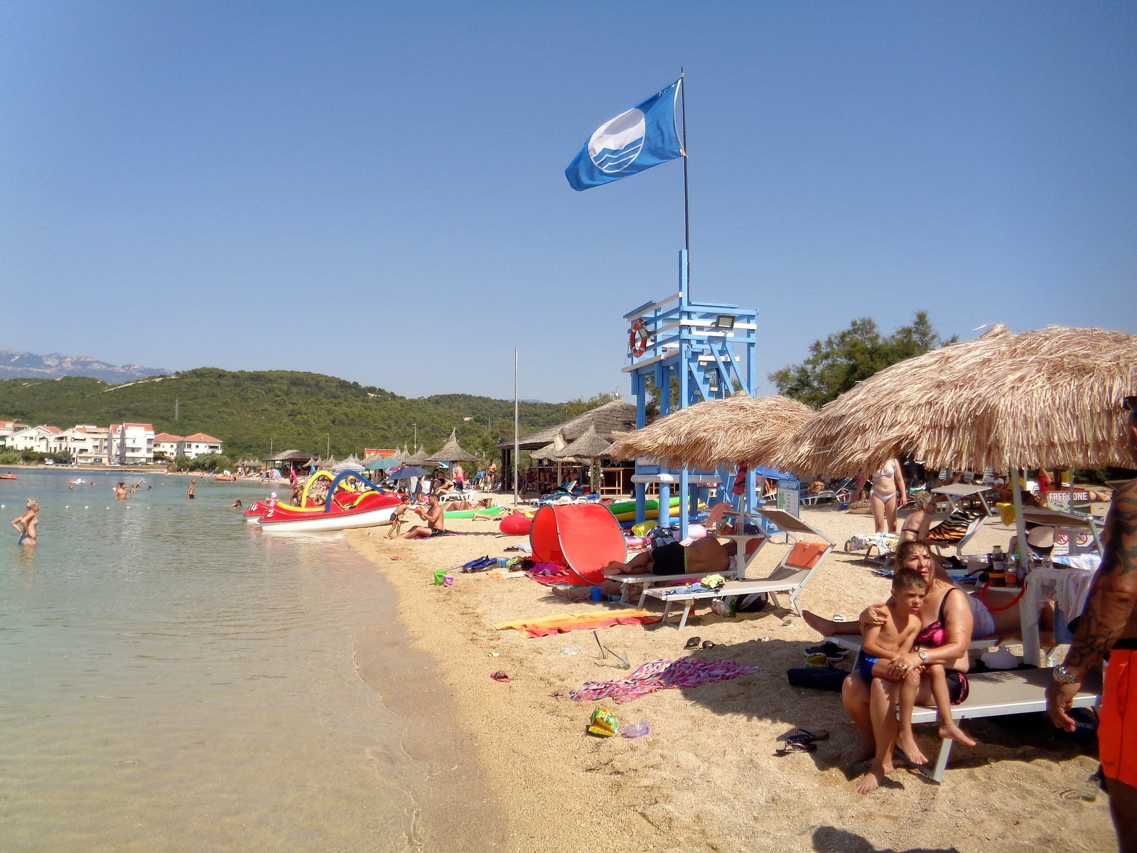 A lifeguard tower and a Blue Flag fly over the sandy Planjka (Trinćel) Beach.