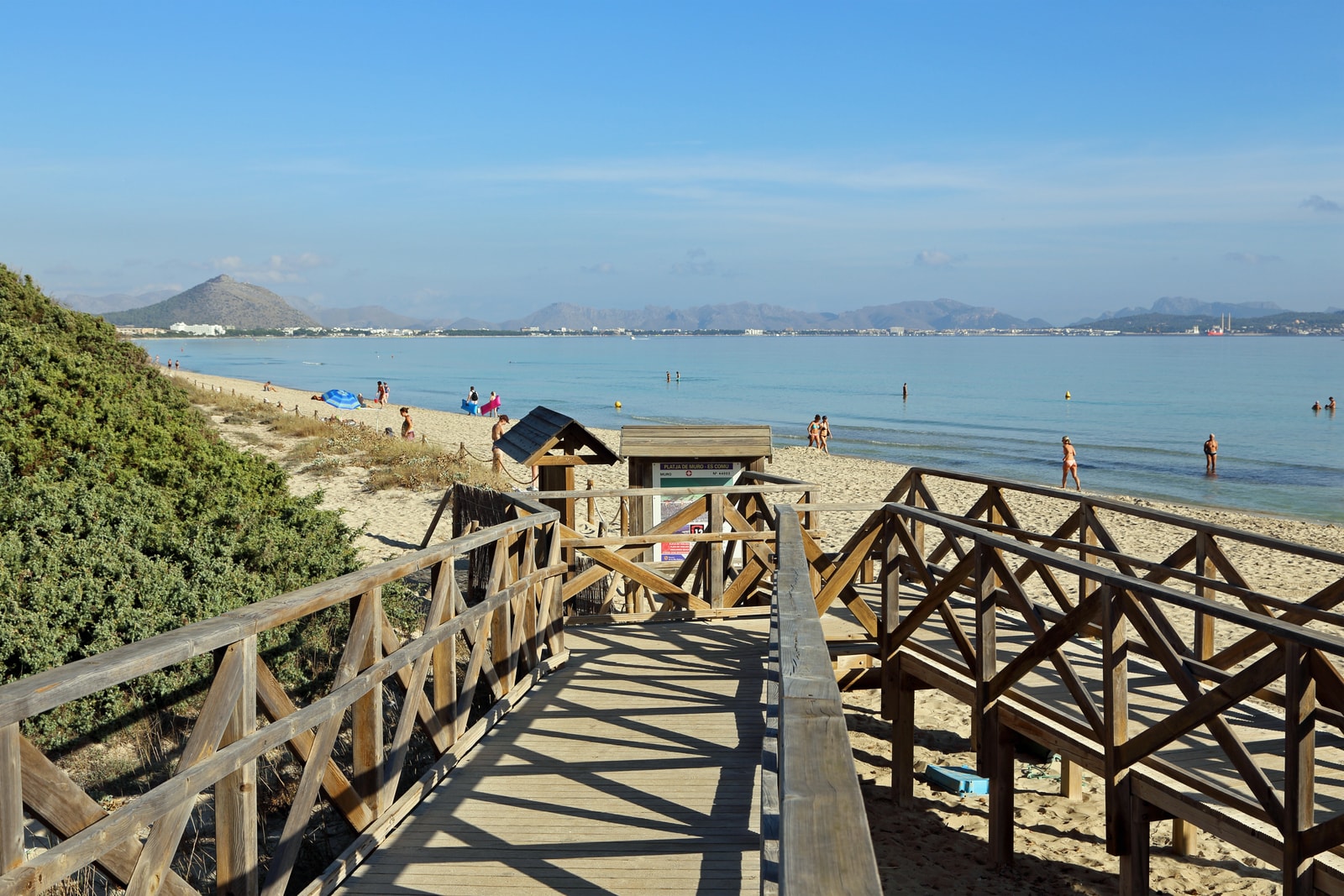A wooden boardwalk leads through low coastal bushes towards a sandy beach.