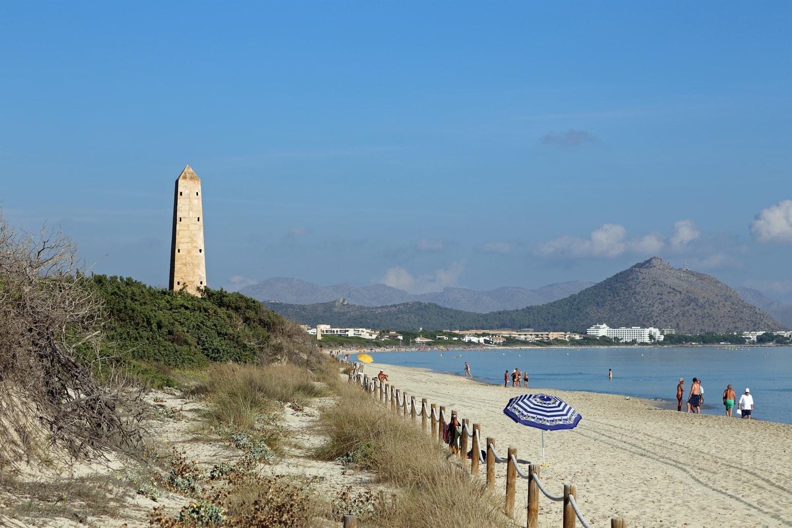 A wooden walkway leads down to a sandy beach with clear blue water, with a small stone tower and mountains visible in the distance.