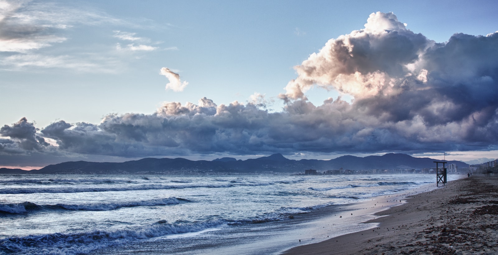 Dramatic clouds loom over a wide sandy beach with waves crashing on the shore and a few people walking in the distance.