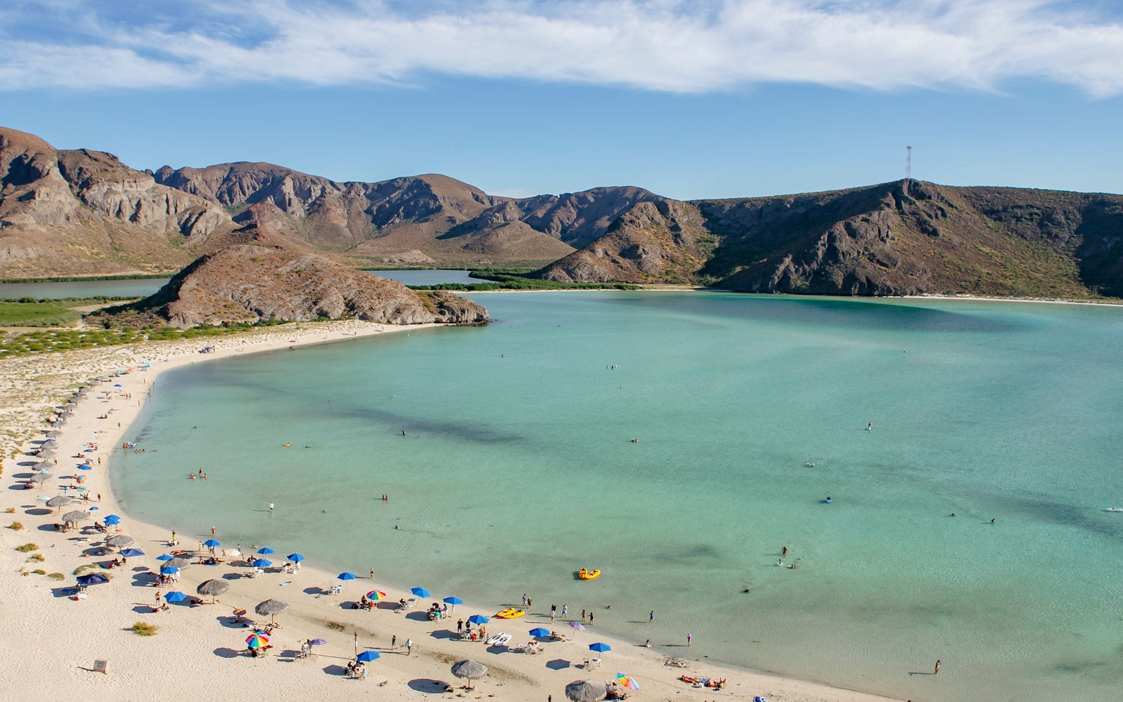 An elevated view of Playa Balandra, showing a crescent of white sand enclosing a shallow, turquoise lagoon, with desert mountains surrounding it.