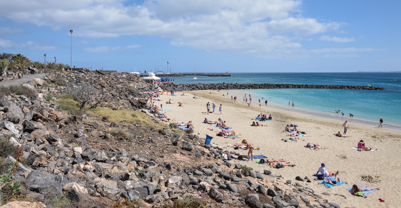 A wide, sandy beach in Playa Blanca bustles with people, with the town's marina visible in the background.