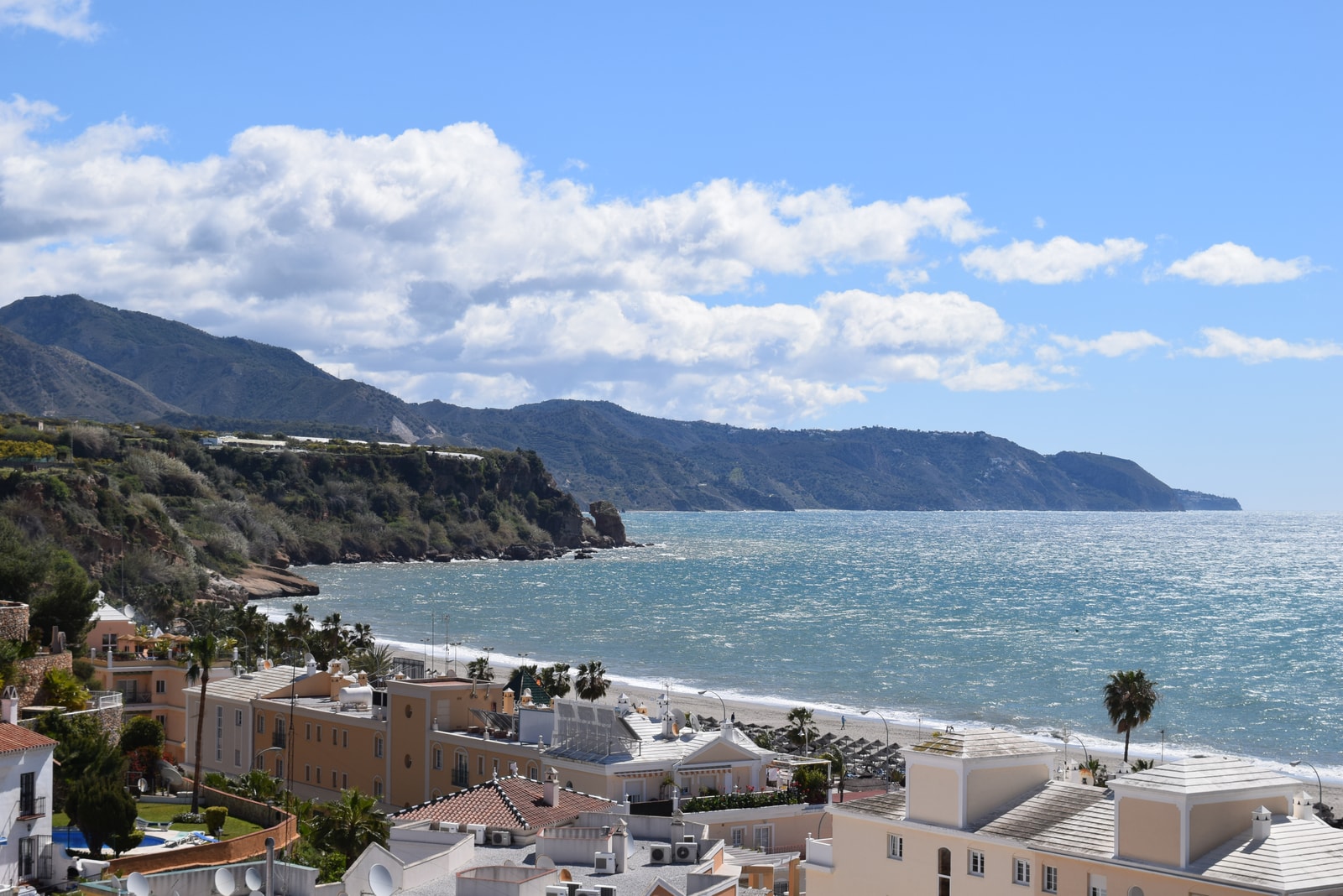 A curving bay with golden sand is backed by palm trees, white resort buildings, and mountains in the distance, showing the expansive Playa de Burriana.