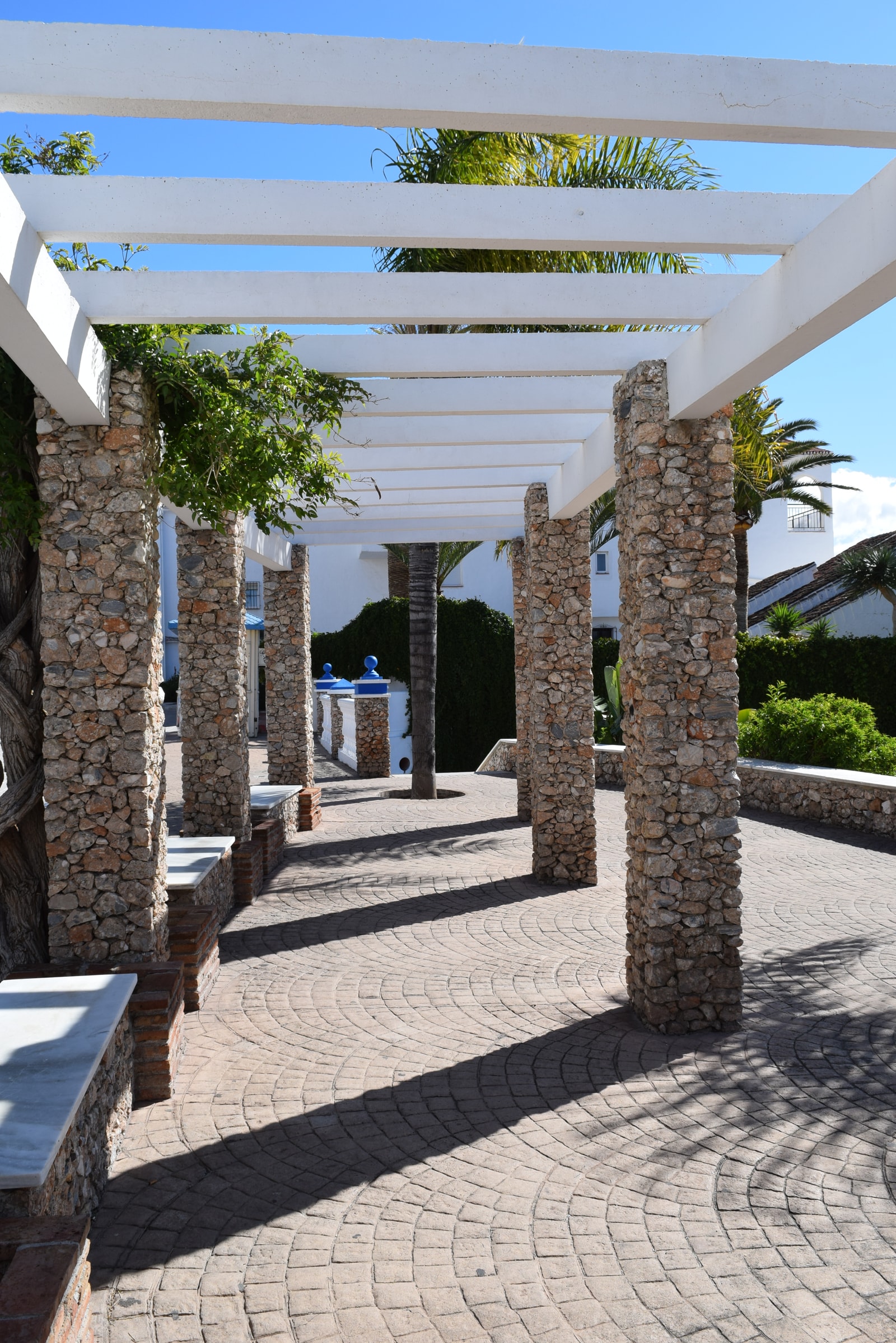 A landscaped sitting area with a white wooden pergola and benches overlooks a lush cove with turquoise water.