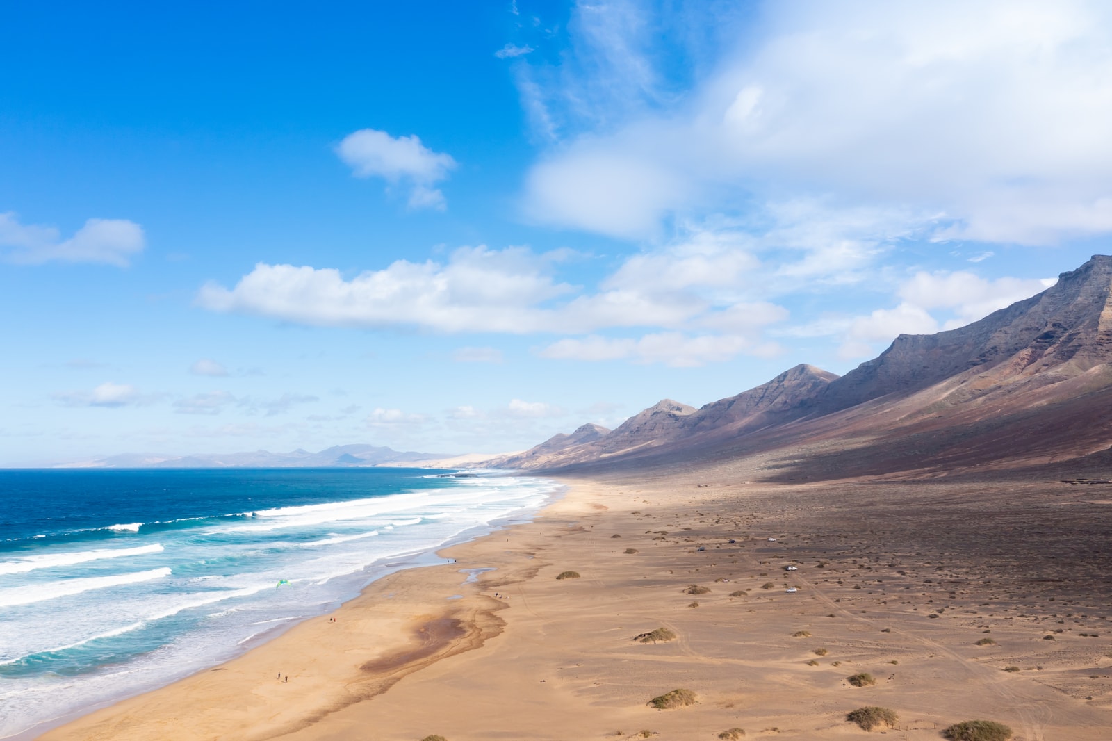 An expansive aerial view of a golden sand beach backed by the imposing, jagged peaks of the Jandia mountains.